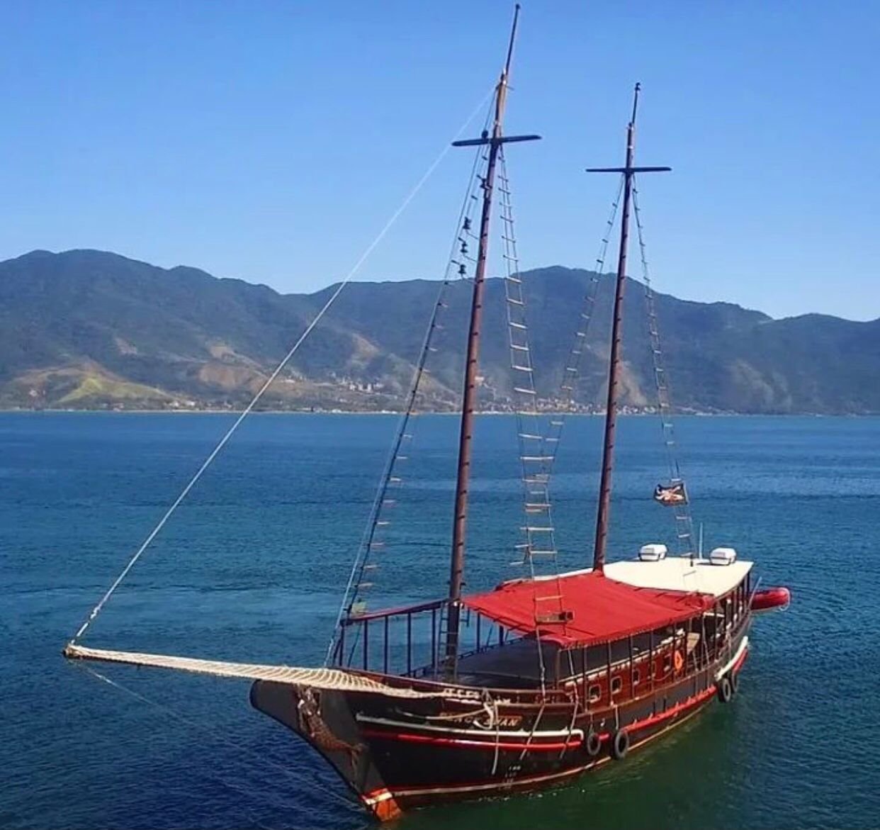 Escuna de dois mastros com toldo vermelho ancorada no mar de Ilhabela, com montanhas ao fundo
